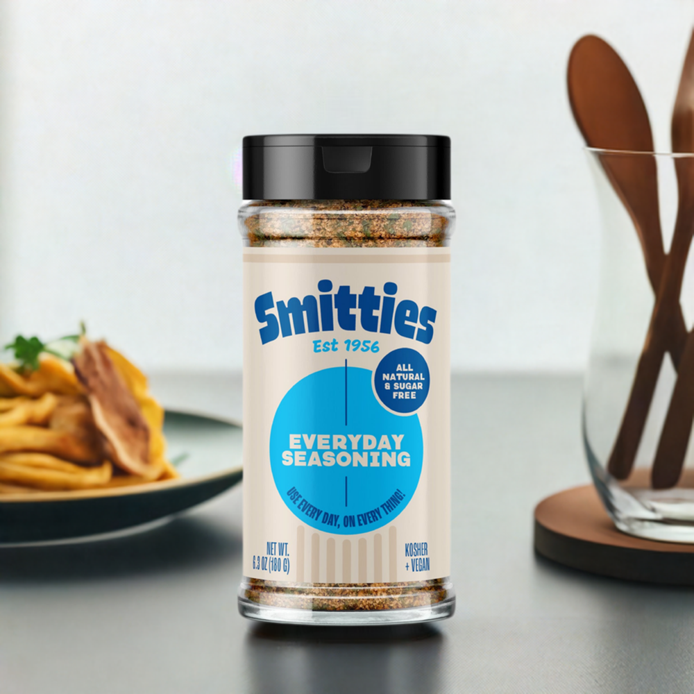A shaker of Smitties Everyday seasoning on a counter, with a plate of french fries and a vase holding wooden spoons in the background