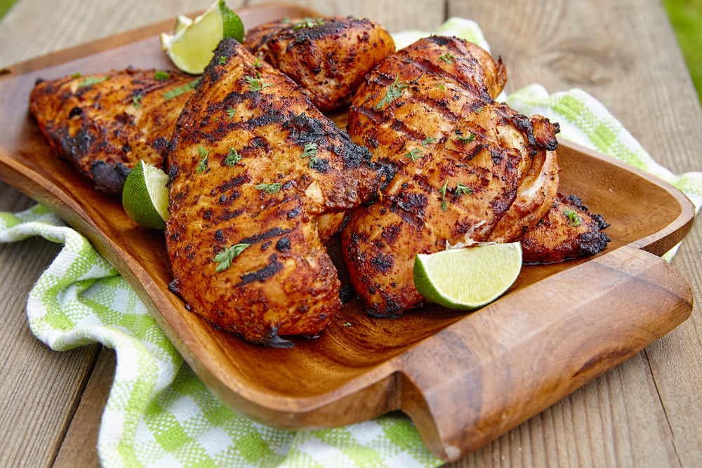 A roasted and grilled chicken on a wooden serving dish, with lime wedges, sitting on a green checkered towel on a wooden table.