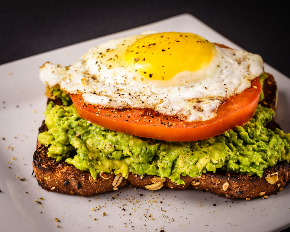 Mashed avocado, a slice of tomato, a fried egg, and sprinkles of spice mix on a piece of whole grain toast.