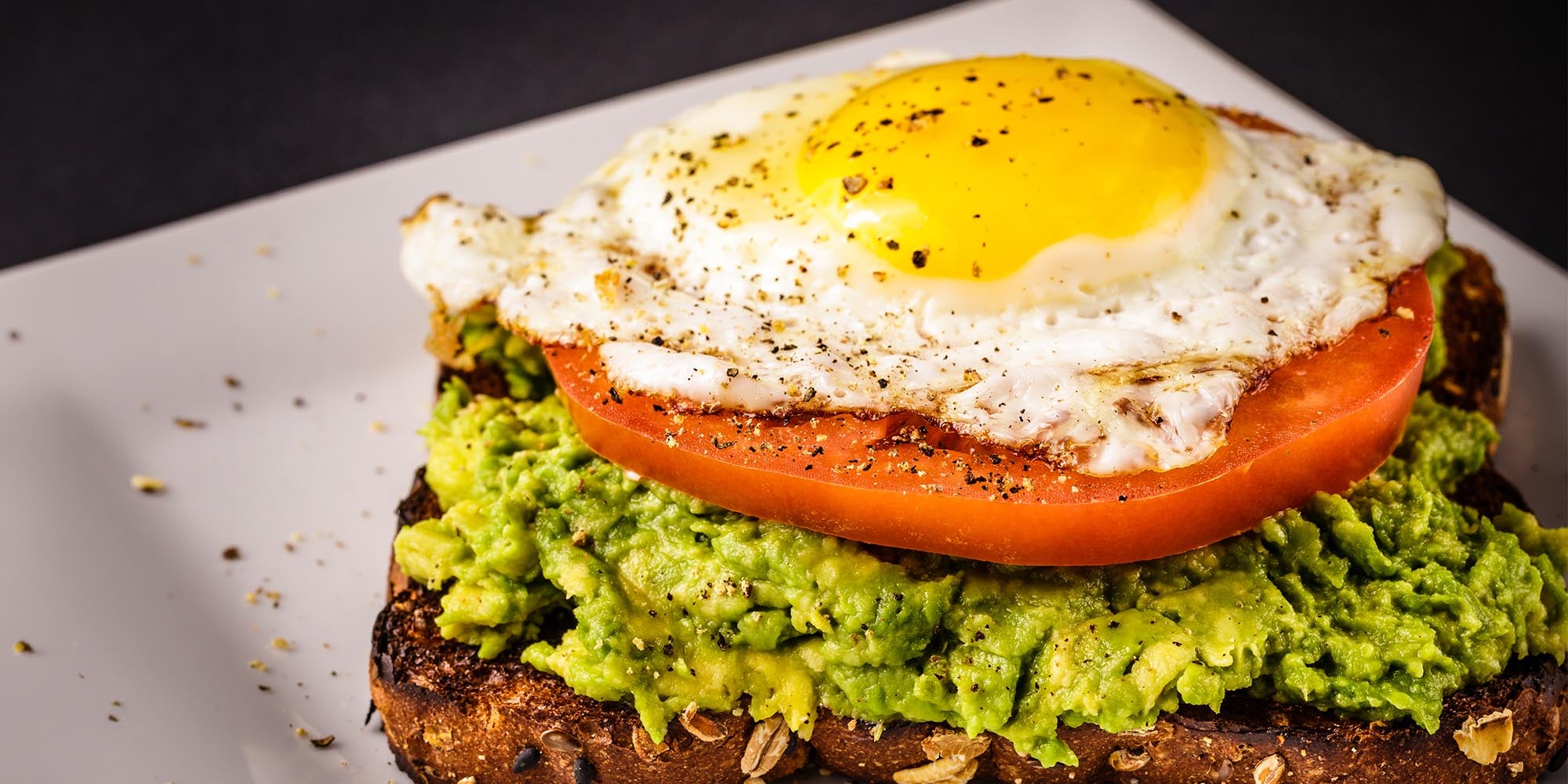 Mashed avocado, a slice of tomato, a fried egg, and sprinkles of spice mix on a piece of whole grain toast.