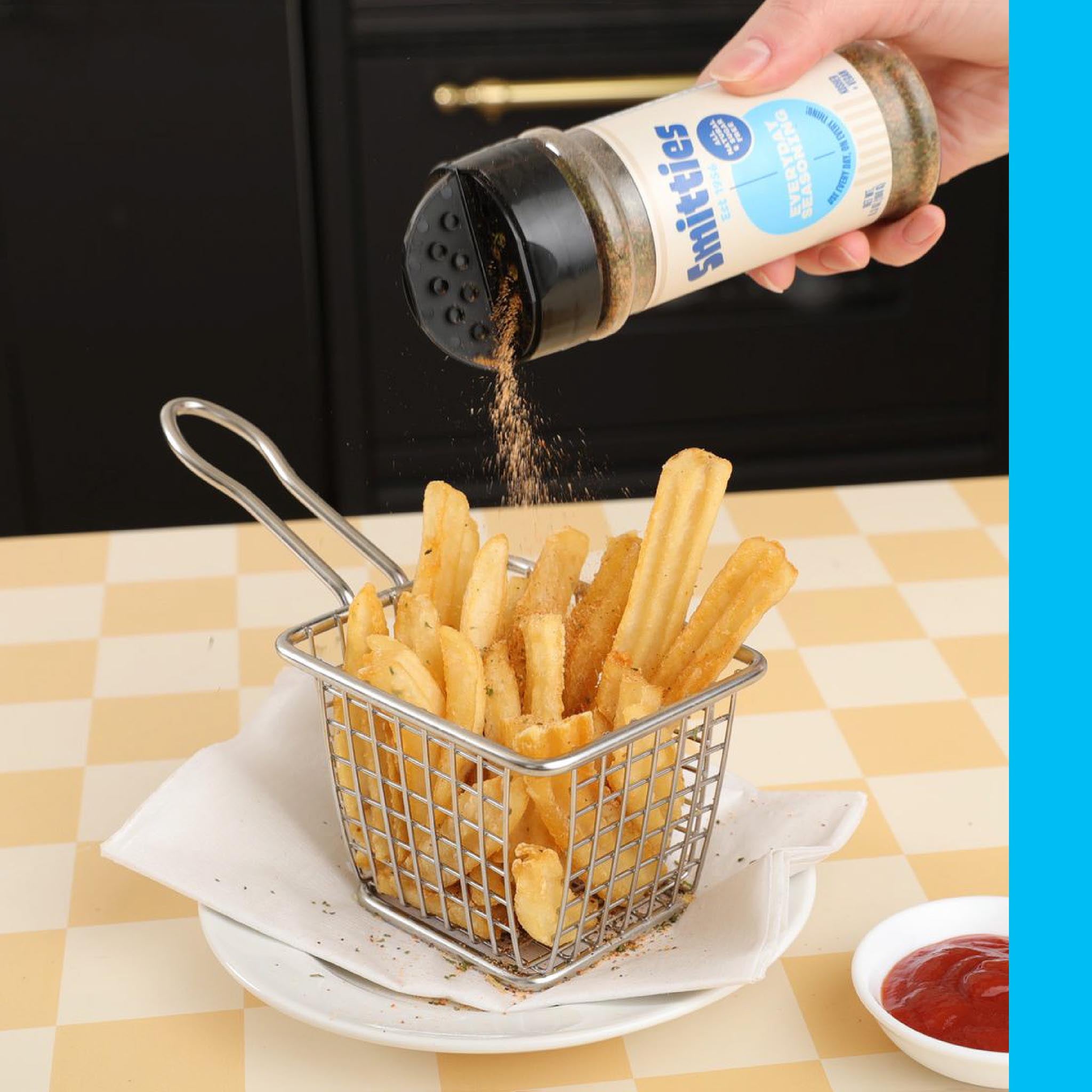 A hand shaking Smitties Everyday Seasoning over a small fryer basket of french fries on a plate, resting on a yellow checked tablecloth next to a bowl of ketchup.