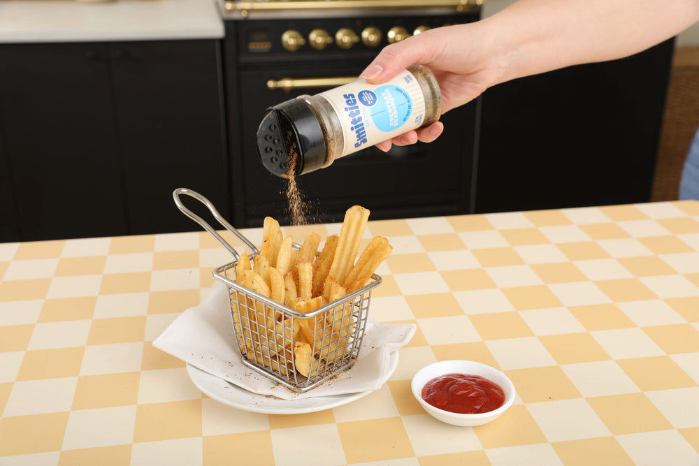 A hand shaking Smitties Everyday Seasoning over a small fryer basket of french fries on a plate, resting on a yellow checked tablecloth next to a bowl of ketchup.
