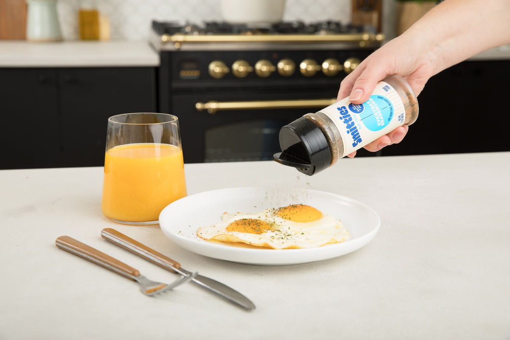 A hand shaking Smitties Everyday Seasoning over a plate of fried eggs, sitting on a white countertop next to a fork and knife, and a glass of orange juice.
