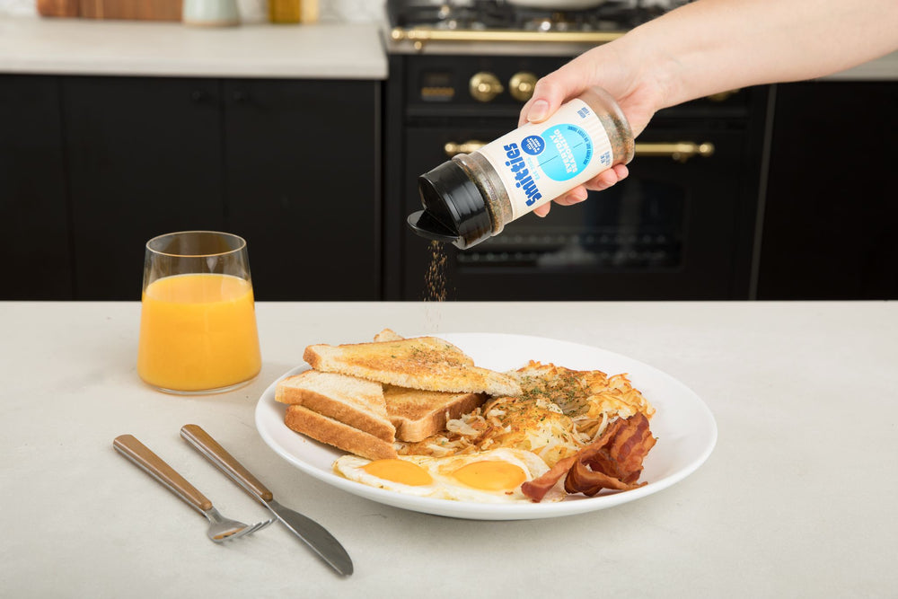 A hand shaking Smitties Everyday Seasoning over a plate of toast, fried eggs, bacon, and hash browns, sitting on a white countertop next to a fork and knife and a glass of orange juice.